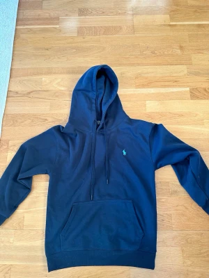 Marinblå hoodie från Polo Ralph Lauren - Snygg marinblå hoodie från Polo Ralph Lauren med klassisk huva och dragsko. Har en känguruficka framtill och ett ljus blått Polo-loggan broderad på bröstet. Storlek S=170
