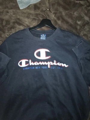 Svart långärmad Champion tröja - Säljer en svart långärmad tröja från Champion med stort tryck på bröstet i vitt, rött och blått. Klassisk rund halsringning och avslappnad passform. Perfekt för dig som gillar streetwear och vill ha en enkel men snygg look.