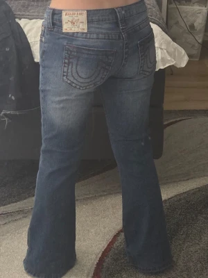 Blå bootcut jeans från True Religion - Säljer ett par klassiska blå bootcut jeans från True Religion med ikoniska kontrastsömmar och broderade bakfickor. Jeansen har låg midja, fem fickor och silverfärgade knappar. Perfekta för dig som gillar en avslappnad men trendig look.