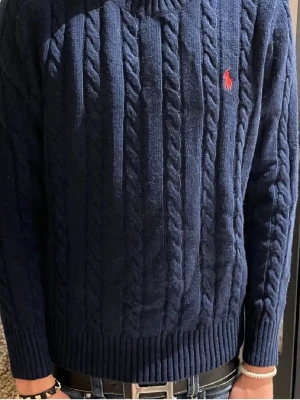 Mörkblå kabelstickad tröja Polo Ralph Lauren - Kabelstickad mörkblå tröja från Polo Ralph Lauren med klassisk röd logga broderad på bröstet. Tröjan har rund halsringning, ribbade muddar och är tillverkad i mjukt material. Perfekt för dig som gillar en stilren och tidlös look. Modellen är 176 väger tröjan är 170 modell 