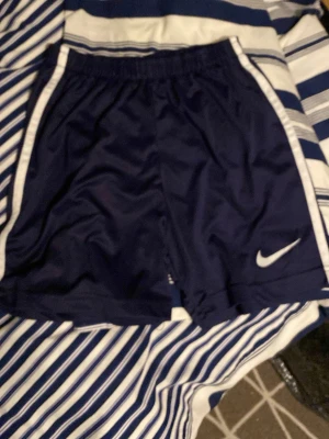 Mörkblå PSG x Nike träningsshorts - Säljer ett par mörkblå träningsshorts från Nike i samarbete med PSG. Shortsen har vita detaljer längs sidorna och en vit Nike-logga på vänster ben. Tillverkade i ett lätt och snabbtorkande material, perfekta för fotboll eller träning.