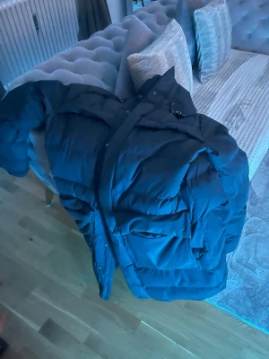 Svart dunjacka från Everest - Säljer en svart dunjacka från Everest med huva och dragsko. Jackan har ett broderat Everest-märke på ärmen och stora fickor framtill. Perfekt för kalla vinterdagar tack vare det tjocka dunfodret och den långa modellen som ger extra värme.