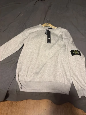 Grå sweatshirt från Stone Island - Säljer en stilren grå sweatshirt från Stone Island med klassisk rund hals och ribbade muddar. Tröjan har den ikoniska Stone Island-loggan på vänster ärm och är tillverkad i mjukt bomullsmaterial. Perfekt för en clean och avslappnad look.
