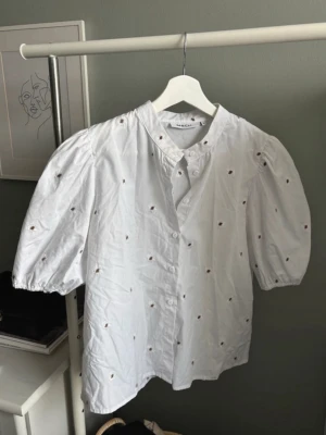 Vit blus med broderade detaljer från Moss Copenhagen  - Söt vit blus från Moss Copenhagen med puffiga trekvartsärmar och små broderade blommor över hela tyget. Blusen har rund halsringning och knäppning framtill. Tillverkad i ett lätt bomullstyg som ger en luftig känsla.