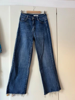 Blåa wide leg jeans med rå kant - Säljer ett par blåa wide leg jeans med hög midja och rå, fransig kant nedtill. Jeansen har klassiska fem fickor och en snygg tvättad look. Perfekta för dig som gillar en avslappnad och trendig stil.