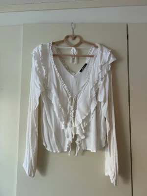 Vit volangblus med spetsdetaljer - Romantisk vit blus från Gina Tricot med långa ärmar, volangdetaljer och spets längs ringningen. Blusen har knytband framtill och lätt struktur i tyget. Perfekt för dig som gillar en bohemisk och söt stil.