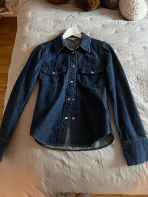 Mörkblå jeansskjorta med knappar - Säljer en mörkblå jeansskjorta med klassisk krage och långa ärmar. Skjortan har två bröstfickor med lock och vita knappar framtill. Perfekt för dig som gillar en avslappnad men snygg stil.