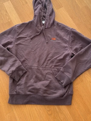 Lila hoodie från New Balance - Säljer en lila hoodie från New Balance med klassisk huva och dragsnören. Hoodien har en magficka framtill och ribbade muddar vid ärmar och nederkant. Liten New Balance-logga i orange på bröstet. Perfekt för en avslappnad och sportig stil.