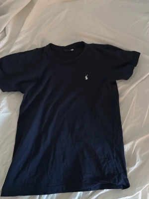 Marinblå t-shirt från Ralph Lauren - Klassisk marinblå t-shirt från Ralph Lauren med den ikoniska vita polospelaren broderad på bröstet. T-shirten har rund hals och korta ärmar, tillverkad i mjuk bomull för en skön känsla. Perfekt basplagg med stilren design. Lite mindre i storleken