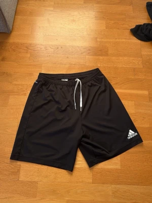 Svarta shorts från Adidas med snörning - Säljer ett par svarta Adidas shorts med vit snörning i midjan och klassisk logga på benet. De har en enkel och sportig design, perfekt för träning eller chill. Tillverkade i ett lätt och bekvämt polyestermaterial.