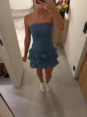 Jeansklänning med volang och axelbandslös - Säljer en snygg axelbandslös jeansklänning med volangkjol och figurnära passform. Klänningen har två lager volanger nedtill och markerade sömmar framtill🫶🏻
