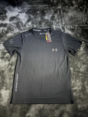 Svart träningströja från Under Armour - Snygg svart träningströja från Under Armour med korta ärmar och rund hals. Tröjan har logga på bröstet och texten UNDER ARMOUR längs sidan. Tillverkad i ett lätt och snabbtorkande funktionsmaterial, perfekt för träning.