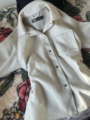 Ljusgrå overshirt från Zara - Stilren ljusgrå overshirt från Zara med klassisk krage, tryckknappar framtill och en stor bröstficka. Overshirten har en mjuk, fleece-liknande yta och raka ärmar.