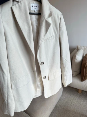Beige kavaj från NA-KD - Stilren beige kavaj från NA-KD med klassisk lite oversized passform, två framfickor (hål i ena) och en bröstficka. Kavajen har två knappar framtill och fyra dekorativa knappar vid ärmsluten. Axelvaddar. Strlk 32. Säljer billigt då den behöver noppras lite och hål i ena fickan💗 Skriv för fler bilder så löser jag det
