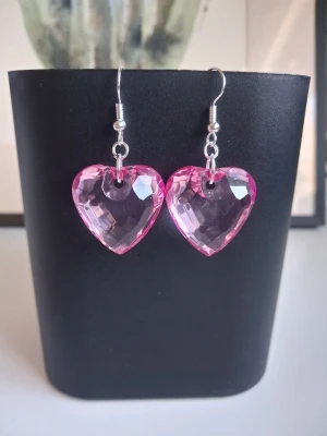 Rosa hjärtformade örhängen i akryl - Säljer ett par söta örhängen med stora, hjärtformade hängen i rosa akryl. De har en glittrande, facettslipad yta som fångar ljuset och ger en extra pop till din look. Krokarna är i metallfärg och örhängena är lätta att bära. Perfekta för dig som gillar statement-accessoarer. Mått 22x22x11cm längd ca 3cm. Öronkroken är nickelsäker och aldrig använda 
