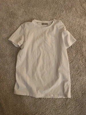 Vit Burberry t-shirt med broderad logga - Säljer en klassisk vit t-shirt från Burberry med broderad B-logga på bröstet. T-shirten har rund halsringning och korta ärmar. Perfekt basplagg med exklusiv känsla och stilren design. Jag tar byten. Den passar S och M
