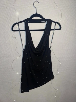 Svart pärlprydd  ZARA - Snygg svart linnetopp med draperad front och silverfärgade pärlor över hela tyget. Toppen är ärmlös och har en djup urringning, perfekt för att sticka ut. Materialet känns mjukt och följsamt, och toppen har en elegant look.