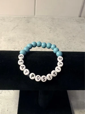 Blått pärlarmband med text - Armband med ljusblå runda pärlor och vita bokstavspärlor som stavar 'STAY POSITIVE'. Elastisk tråd gör det enkelt att ta på och av. Perfekt accessoar för dig som gillar positiva budskap och färgglada detaljer.