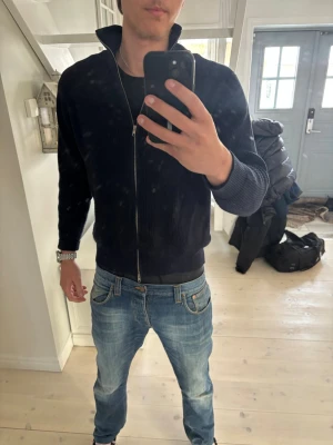 Maison Margiela Zip Tröja - Säljer en mörkblå Maison Margiela ziptröja med hög krage och ribbade muddar. Tröjan har hel dragkedja framtill och är tillverkad i ett mjukt, stickat material. Perfekt för dig som gillar en clean och stilren look.
