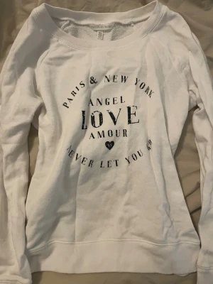 Vit sweatshirt från Victoria's Secret - Vit sweatshirt från Victoria's Secret med svart texttryck på bröstet där det står bland annat 'Paris & New York', 'Angel', 'Love', 'Amour' och 'Never let you go'. Tröjan har rund halsringning och är tillverkad i mjuk bomull.