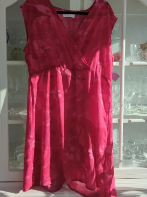 Rosa omlottklänning från Sansa - Snygg rosa omlottklänning från Sansa Collection i lätt och luftig polyester. Klänningen har ett subtilt blommigt mönster, är ärmlös och har en v-ringad omlottdesign som ger en feminin siluett. Perfekt för varma dagar och somriga tillfällen.