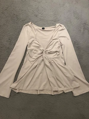 Beige långärmad babydoll topp med twistdetalj - Trendig beige topp med långa ärmar och snygg twistdetalj framtill. Toppen har en lätt utsvängd passform och djup v-ringning som ger en feminin look. Perfekt att styla med jeans eller kjol för en chic vibe.