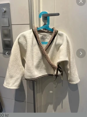 Vit omlottkofta från Bissen storlek 104cm/4 år - Säljer en vit omlottkofta från Hissning med beige och bruna detaljer längs kanterna. Koftan har långa ärmar, knytband i sidan och är gjord i ett mjukt, fleece-liknande material. Perfekt för lager på lager och en chill look.