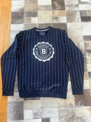 Mörkblå randig sweatshirt Bondelid - Mörkblå sweatshirt från Bondelid med vita vertikala ränder och ett stort tryck med texten 'Bondelid Nineteen Fifty Six' på bröstet. Tröjan har rund halsringning och ribbade muddar vid ärmslut och nederkant. Perfekt för en avslappnad och stilren look.