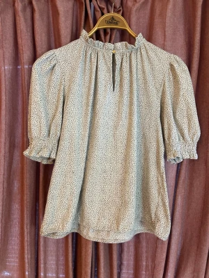 Beige prickig blus med puffärm - Söt vit blus med små svarta prickar och puffiga ärmar. Blusen har volang vid halsringningen och en liten öppning framtill med knapp. Materialet är lätt och luftigt, perfekt för en trendig och avslappnad look.