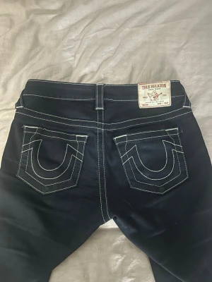 True Religion Becca Mid Rise Bootcut Jeans - Mörkblå jeans från True Religion, modell Becca Mid Rise Bootcut. Snygga kontrastsömmar och ikoniska fickdetaljer bak. Klassisk bootcut-passform och mellanmidja, perfekta för en avslappnad men trendig look.