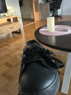 Svarta låga sneakers i skinnimitation - Stilrena svarta sneakers med lågt skaft och matchande svarta skosnören. Skorna har en clean look med ovandel i skinnimitation och svarta metalldetaljer vid snörningen. Perfekta för dig som gillar minimalistisk och tidlös stil.