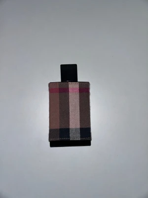 Burberry - Säljer en Burberry parfymflaska med det klassiska rutiga tygfodralet i beige, brunt, svart och rött. Flaskan har en rektangulär form och svart lock. Perfekt för dig som gillar stilrena och ikoniska designer.