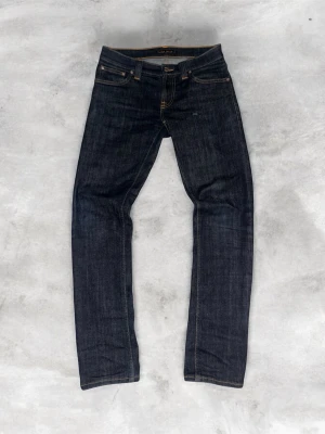 Mörkblå jeans från Nudie Jeans - Säljer ett par mörkblå jeans från Nudie Jeans med klassisk femficksmodell och snygga kontrastsömmar. Jeansen har rak passform och är gjorda i kraftig denim med läderpatch bak i midjan. Perfekta för en clean och stilren look.