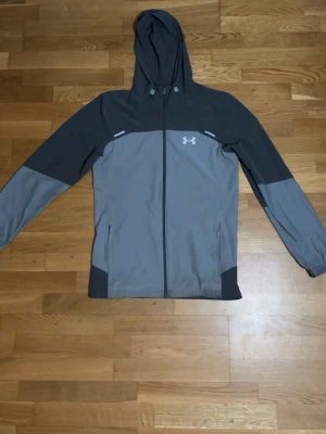 Grå vindjacka från Under Armour - Snygg tvåfärgad vindjacka från Under Armour i ljusgrått och mörkgrått. Jackan har huva, hel dragkedja framtill, två sidofickor och en vit logga på bröstet. Tillverkad i lätt och vindtätt material, perfekt för blåsiga dagar.