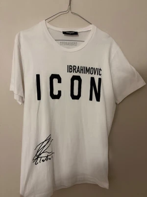 Vit ICON t-shirt från Dsquared2 - Vit t-shirt från Dsquared2 med svart text 'ICON IBRAHIMOVIC' framtill och en signaturdetalj. Baksidan har ett tryck med porträtt och citat. Klassisk rund hals och korta ärmar. Perfekt för dig som gillar statement-plagg och sportig stil.