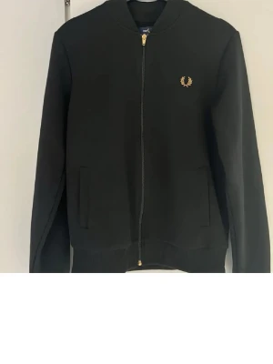 Svart zip hoodie från Fred Perry - Snygg svart hoodie från Fred Perry med dragkedja framtill och två fickor. Klassisk design med guldfärgad logga broderad på bröstet. Tillverkad i mjukt material. 
