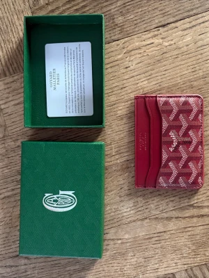 Vinröd Goyard Plånbok - Snygg röd kortplånbok från Goyard med det klassiska vita och gråa mönstret. Plånboken har flera kortfack och är tillverkad i coated canvas och läder. Perfekt för dig som vill ha något stilrent och lyxigt i fickan. 