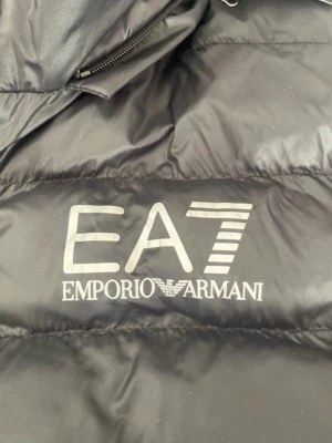 Svart pufferjacka från EA7 Armani - Snygg svart pufferjacka från EA7 Emporio Armani med stor logga på bröstet. Jackan har huva, dragkedja framtill och quiltad design. Perfekt för kalla dagar och ger en sportig, trendig look. Materialet är glansigt och känns lätt men ändå varmt. Är i nyskick.
