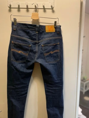 Mörkblå skinny jeans från Nudie - Säljer ett par mörkblå skinny jeans från Nudie Jeans med klassiska orange sömmar och broderade bakfickor. Jeansen har fem fickor, bälteshällor och är tillverkade i stretchig denim för en bekväm passform.
