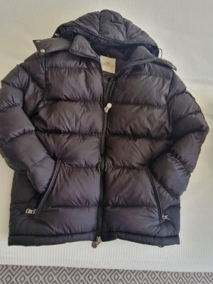 Svart dunjacka från Moncler - Snygg svart dunjacka från Moncler med huva och dragkedja framtill. Jackan har två sidofickor med dragkedja och klassisk quiltad design. Insidan har Monclers ikoniska serietidningsetikett. Perfekt för kalla vinterdagar.