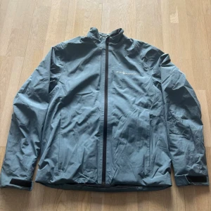 Grå vindjacka från Sail racing - Snygg grå vindjacka från Sail Racing med hel dragkedja framtill och hög krage. Jackan har justerbara muddar med kardborre och diskreta loggor på bröstet och ärmen. Tillverkad i lätt och vindtätt material, perfekt för blåsiga dagar.