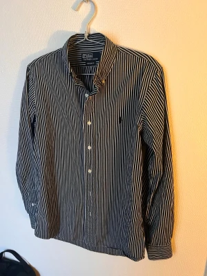 Randig skjorta från Polo Ralph Lauren - Snygg svartvit randig skjorta från Polo Ralph Lauren i modellen Custom Fit. Skjortan har klassisk krage, knäppning framtill och lång ärm. Perfekt för dig som gillar stilrena plagg med ikonisk logga på bröstet.