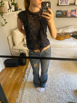 Mörkblå spetstopp  - Säljer en mörkblå blus i spets med volangdetaljer längs framsidan och ärmarna. Toppen är ärmlös och är genomskinlig. Perfekt att styla med jeans eller kjol.