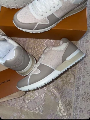 Beige Louis Vuitton sneakers - Stilrena sneakers från Louis Vuitton i beige och vitt med klassisk LV-logga på sidan. Skorna har en kombination av mocka och textil, vita snören och en vit sula med beige detaljer. Perfekta för dig som gillar exklusiv och trendig streetstyle.