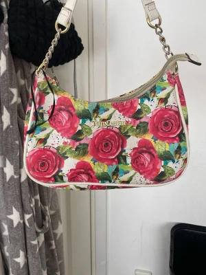 Juicy Couture rosig axelremsväska - Juicy Couture handväska med färgglatt ros-mönster i rosa, gröntg och vitt. Väskan har guldfärgade detaljer, kedjerem och dragkedja upptill. Väskan är köpt i USA för 80 dollar som motsvarar 741kr ish. Väskan är använd ett fåtal gånger.