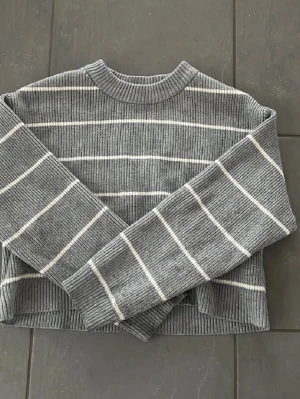 Grå ribbstickad tröja med ränder - En grå ribbstickad tröja med vita horisontella ränder och rund halsringning. Tröjan har långa ärmar och en något boxig, kortare passform. Materialet ser ut att vara mjukt och bekvämt, perfekt för lager-på-lager under kyliga dagar.