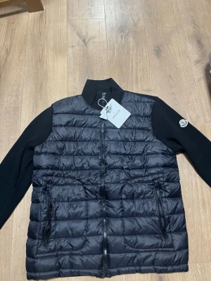 Svart cardiganjacka från Moncler - Snygg svart cardiganjacka från Moncler med quiltad front och stickade ärmar och rygg. Jackan har dragkedja framtill, två fickor med dragkedja och Moncler-logga på ärmen. Perfekt för lager-på-lager och streetwear-vibe.