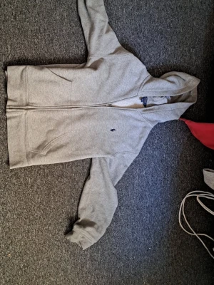Grå hoodie från Polo Ralph Lauren - Snygg grå hoodie från Polo Ralph Lauren med dragkedja framtill och klassisk huva. Tröjan har två fickor och den ikoniska lilla loggan broderad på bröstet. Tillverkad i mjukt bomullsmaterial som känns skönt mot huden. Perfekt för en avslappnad och stilren look.