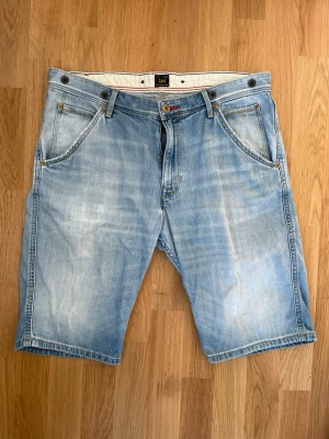 Ljusblå jeansshorts från Lee - Säljer ett par ljusblå jeansshorts från Lee med klassisk femficksdesign och knappgylf. Shortsen har normal passform och är tillverkade i bomull. Perfekta för sommaren och har en avslappnad look med slitningar och tvättade detaljer. Storlek W33