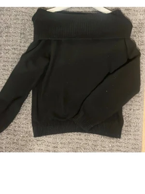 Svart off shoulder stickad tröja Storlek 158/164 - Säljer en svart stickad tröja med bred ribbad kant upptill som kan bäras off shoulder. Tröjan har långa ärmar och ribbade muddar vid ärmslut och nederkant. Perfekt för dig som gillar en enkel men trendig look. Materialet känns mjukt och stretchigt. Storlek 158/164